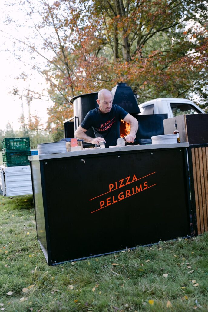 Pizzabakker van Pizza Pelgrims aan de houtoven achter de zwarte stand met logo